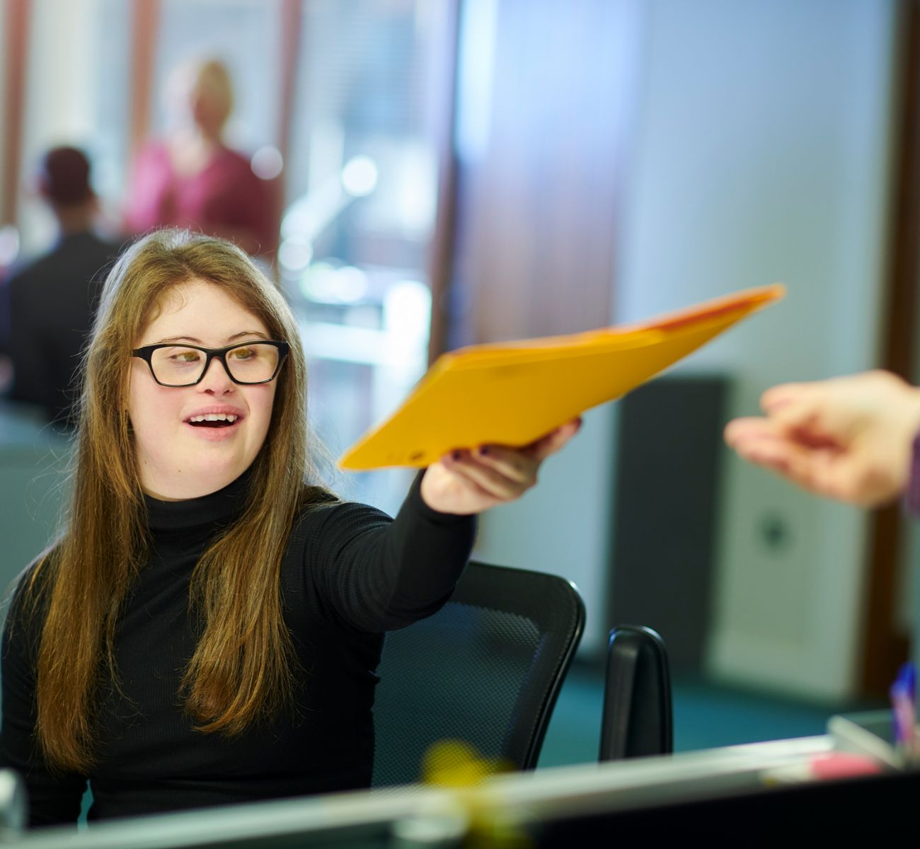 A young woman in a black turtleneck offers a yellow folder to someone in an office setting.