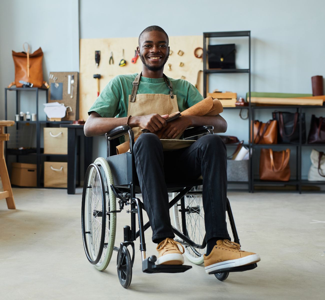Man in a wheelchair smiling, wearing an apron, sits in a workshop with leather goods on shelves and a workbench in the background.