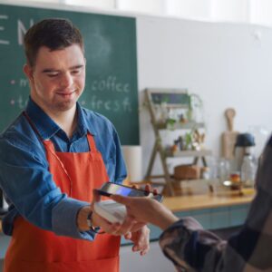 A barista in an orange apron holds a payment terminal while a customer pays with a smartphone in a cozy cafe setting.