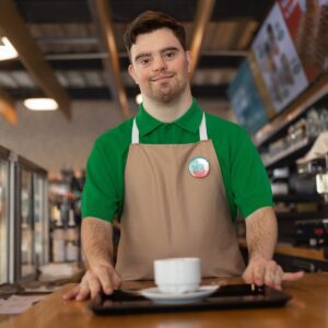 A barista in a green shirt and beige apron stands behind a counter, holding a tray with a cup, in a coffee shop setting.