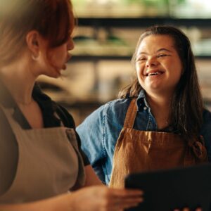 Two women wearing aprons are smiling and talking in a warmly lit room. One holds a tablet.