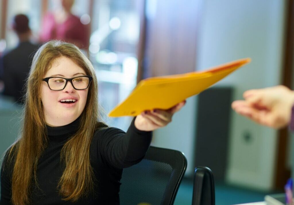 A young woman in a black turtleneck offers a yellow folder to someone in an office setting.