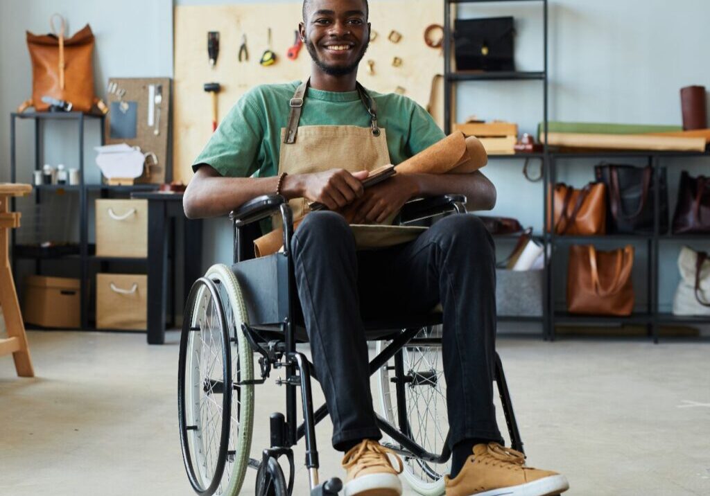 Man in a wheelchair smiling, wearing an apron, sits in a workshop with leather goods on shelves and a workbench in the background.
